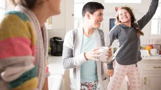 A woman dancing in the kitchen with her daughters and drinking coffee.