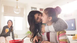 A family of three women are in the kitchen, one daughter hugging the youngest while she flips pancakes.