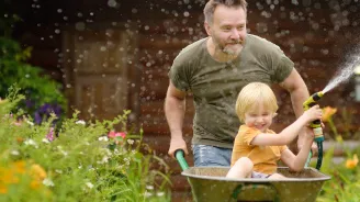 A Father and Son Spray a Garden Hose During a Wheelbarrow Ride