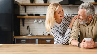 An older couple smiling at each other while in a kitchen.