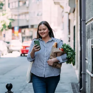 A Woman Walks Down a City Street Reading Her Cell Phone While Carrying Her Groceries