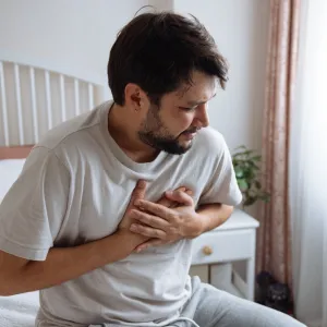 A Man Holds his Hands to his Chest While Sitting on his Bed.