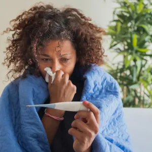 A Woman Blows Her Nose While Sitting on Her Couch at Home Reading a Thermometer