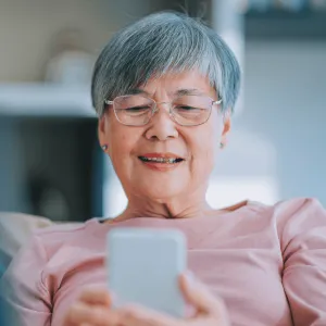 A Senior Woman Sits on the Couch at Home and Scrolls on Her Cell Phone