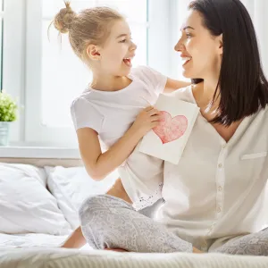 Mother and daughter sitting on a bed, holding each other with a card with a heart on it in the daughter's hands.