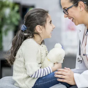 A child sitting on an exam table with her doctor, who has her hand on her knee and is smiling at her.