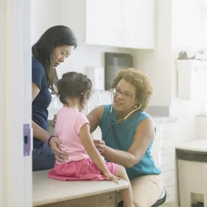 A female AHS doctor listens to a little girl's heart in the exam room while her mother comforts her