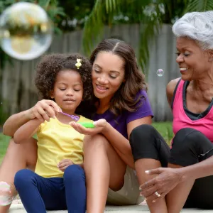 A grandmother, mother, and daughter play with bubbles outside.
