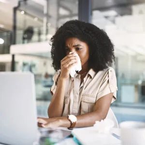 A woman blowing her nose, looking at a laptop.