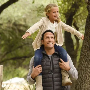 A Caucasian father carries his daughter on his shoulders while on a hike.