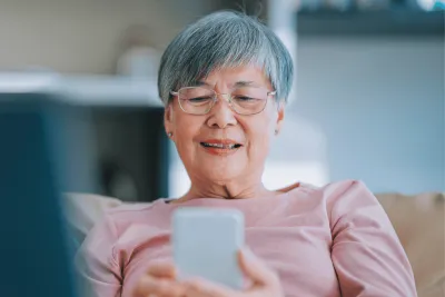 A Senior Woman Sits on the Couch at Home and Scrolls on Her Cell Phone