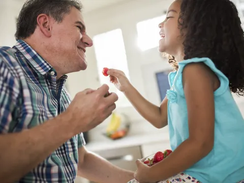 A grandfather and his granddaughter laugh as they eat strawberries in the kitchen.