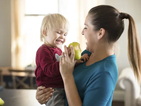 A Caucasian mom feeds her son a pear in the kitchen.