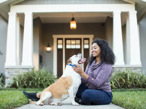 A young woman sits outside of a nice home, playing with her bulldog
