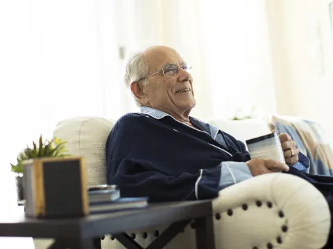 An elderly Caucasian man sits on the sofa with a mug of coffee