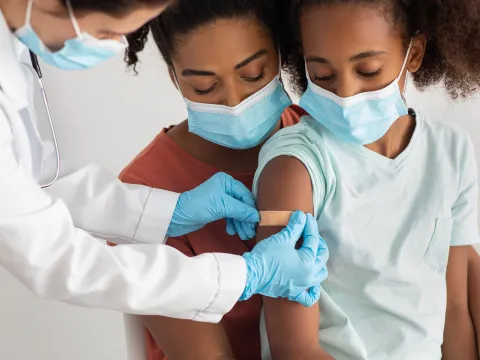 A Mother Comforts Her Daughter While a Physician Puts a Bandaid on an Injection Site in the Upper Arm.