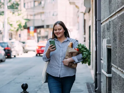 A Woman Walks Down a City Street Reading Her Cell Phone While Carrying Her Groceries