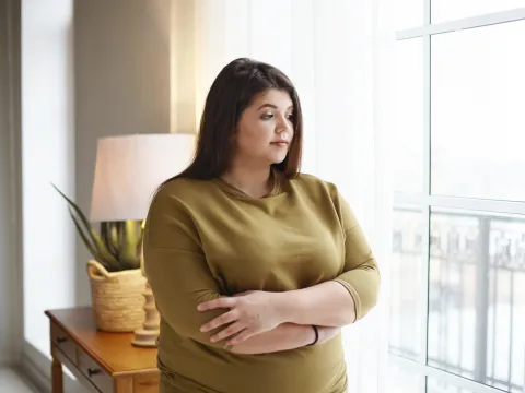 A Woman Holds Her Arms as She Looks Out the Window of Her Home