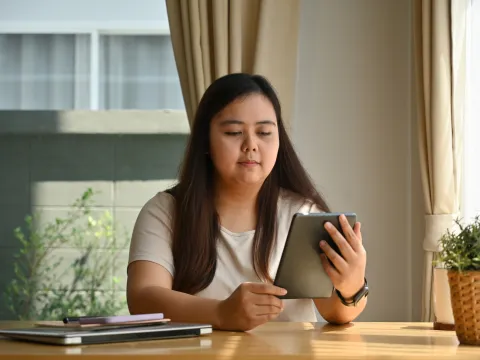 A Woman Reads a Tablet While Sitting at a Table at Home