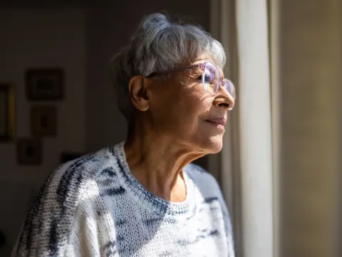A Senior Woman Looks Out The Window of Her Home with a Smile