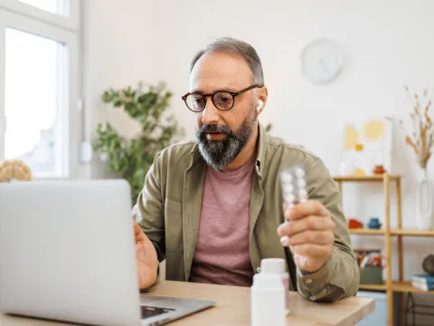 Man attending a virtual care appointment at home on laptop holding medication.