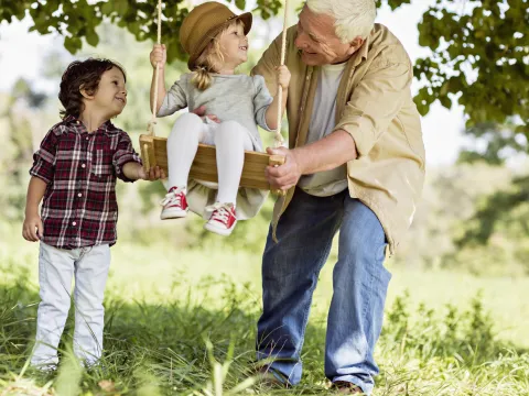 A grandfather with his two grandkids, one sitting on a wood swing, the other helping their grandfather push the swing.