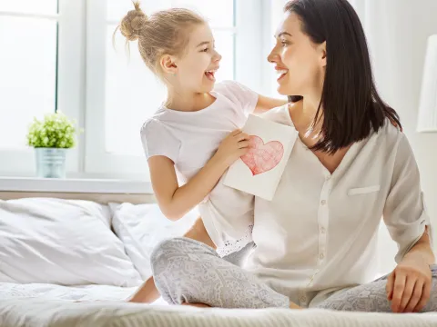 Mother and daughter sitting on a bed, holding each other with a card with a heart on it in the daughter's hands.