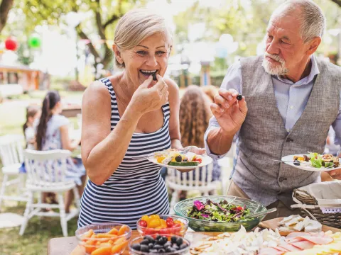 A senior couple at an outdoor party enjoys a heart healthy lunch together. They are standing eating over a buffet table.