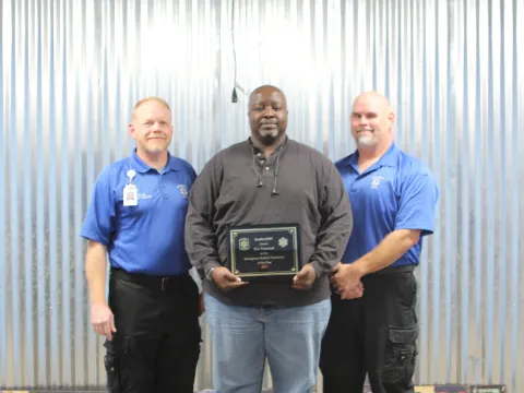 Three men standing next to each other, the man in the middle is holding an award.