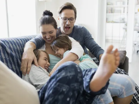 A Caucasian family laughs as they pile on the couch together.