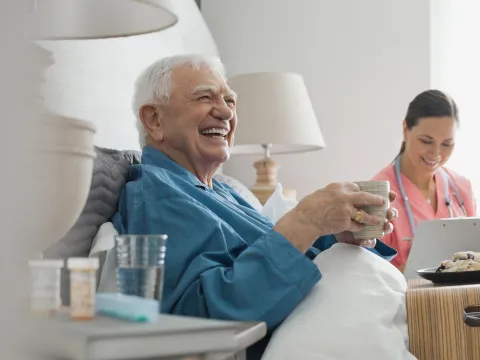 An elderly man seating on his bed in company of a nurse