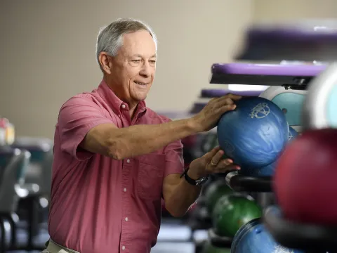 An adult man crouches to choose a bowling ball at the bowling alley.