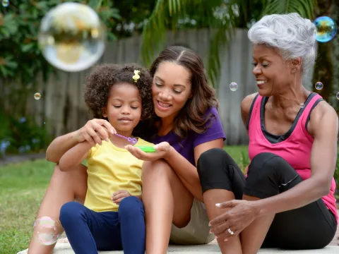 A grandmother, mother, and daughter play with bubbles outside.