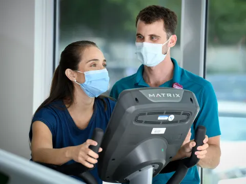 Woman riding a stationary bike while speaking with a trainer at the AdventHealth Health Park gym.