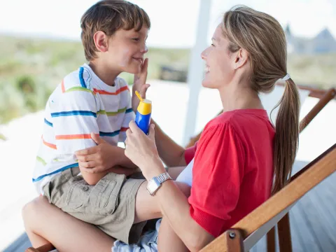 A mom with her son in her lap sitting outside in a char, applying sunscreen to his face