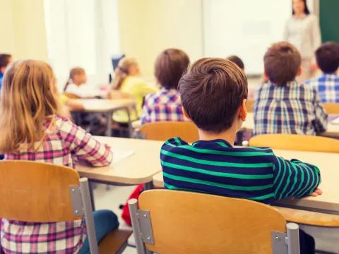 Image of the back of a classroom with several children sitting at their desks facing their teacher and a blackboard.