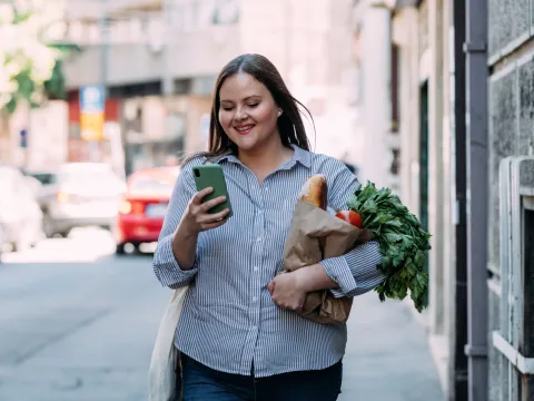 Smiling woman walking and looking at her phone while carrying a bag of groceries.