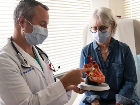Doctor showing a woman patient a heart model while both wear masks.