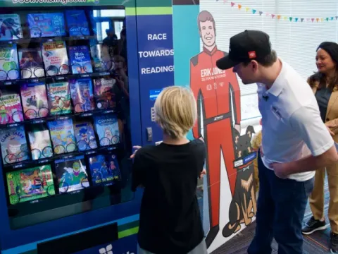 Erik Jones, driver of the No. 43 AdventHealth Toyota Camry XSE, helps a Chisholm Elementary School student use the new book vending machine for the first time.
