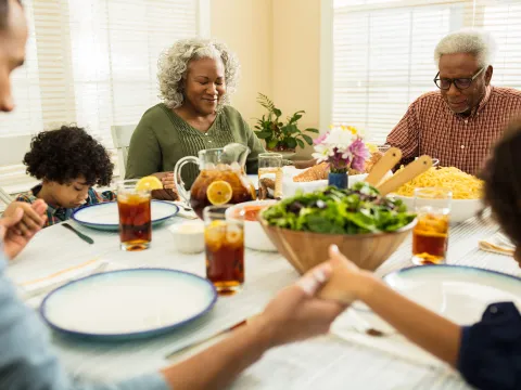 A family praying together while holding hands before eating lunch at home.