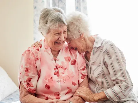 Two older women laugh together while sitting on a bed.