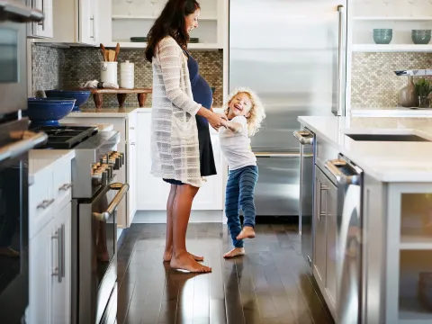 A pregnant mom and her daughter laugh in the kitchen.