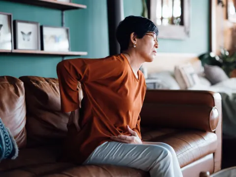 A woman holding her back in pain while sitting on a sofa.