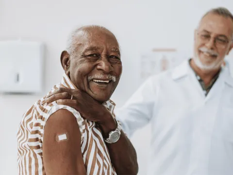A senior man smiles and shows his arm with bandage after receiving a vaccine in the foreground while a doctor smiles in the background.