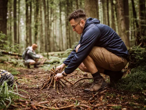 Man building a campfire in a forest while a woman collects timber in the background.