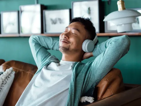 A man sits on a couch at home with listening to music with headphones with his eyes closed, smiling, and hands behind his head.