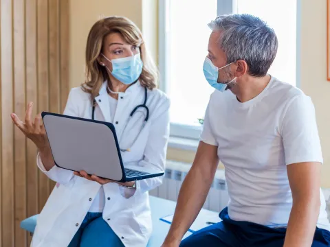 Female doctor talking with male patient holding laptop, both masked