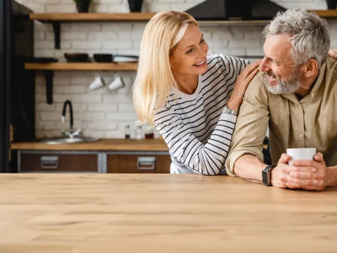 An older couple smiling at each other while in a kitchen.