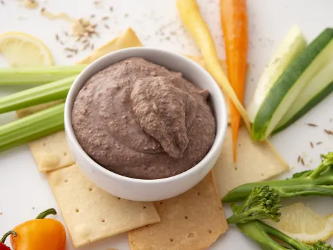 White bowl of black bean hummus surrounded by vegetable sticks and crackers.