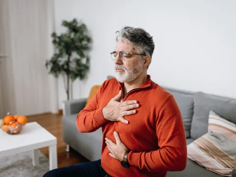 Senior man sitting on a couch at home placing hands on chest and abdomen.